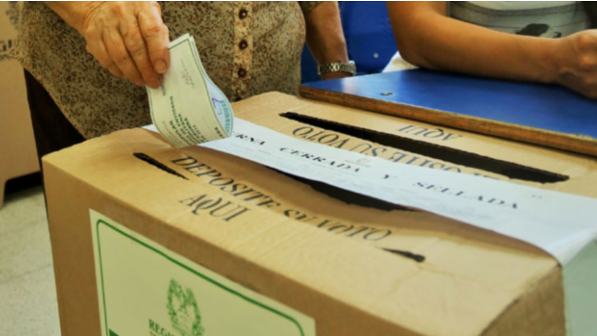 Una caja que funge como urna de votación con una ranura encima por la cual una mano va a introducir un papel de voto.