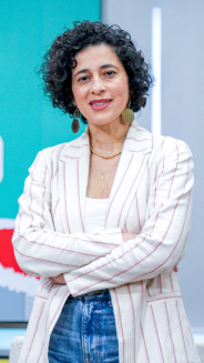 Magda Páez-Periodista. Mujer en plano americano con los brazos cruzados, pelo negro. ondulado y corto. Viste con un blazer blanco con rayas y un jean.