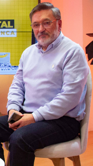 Foto de Carlos Chica, sentado en una silla con camisa y saco color lila; y un jeans